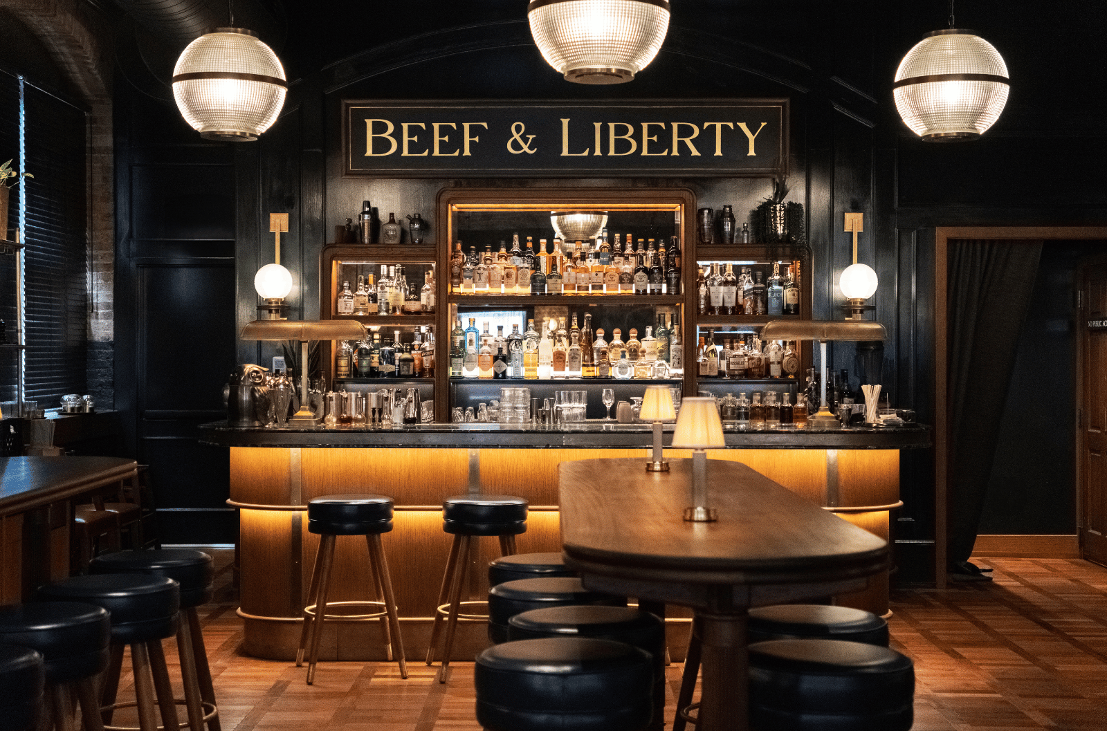 "Beef & Liberty" bar area featuring blue stools, a marble counter, wood tables, small lamp lights, and a backdrop of spirits and glassware.