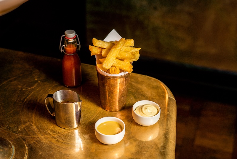 A pot of beef-dripping chips on a golden table, served in a brass pot with homemade condiments accompanying it.