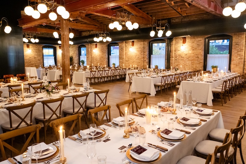 A wide shot of the Beef and Liberty bar, which is a large room with big windows, allowing daylight to pour through. It is set up for a wedding with 6 long tables visible, all covered by white table cloths and table decor, including candles.