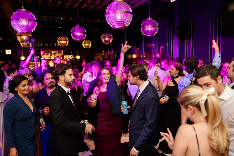A room full of smartly dressed people, having fun, dancing and drinking cocktails, under disco balls and purple lights.