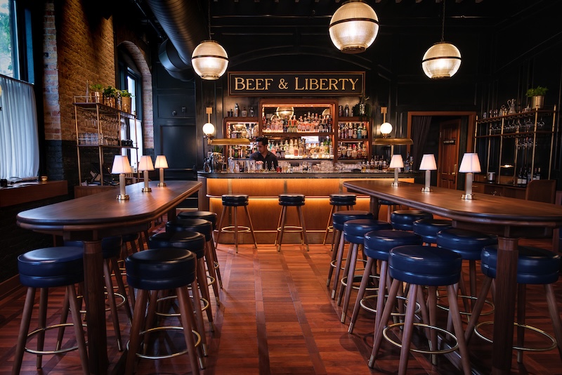 A bar facing view of the Beef and Liberty Bar, with three bar stools visible lined up along the bar. In the foreground are two tables and stools, each table has 8 spaces available.