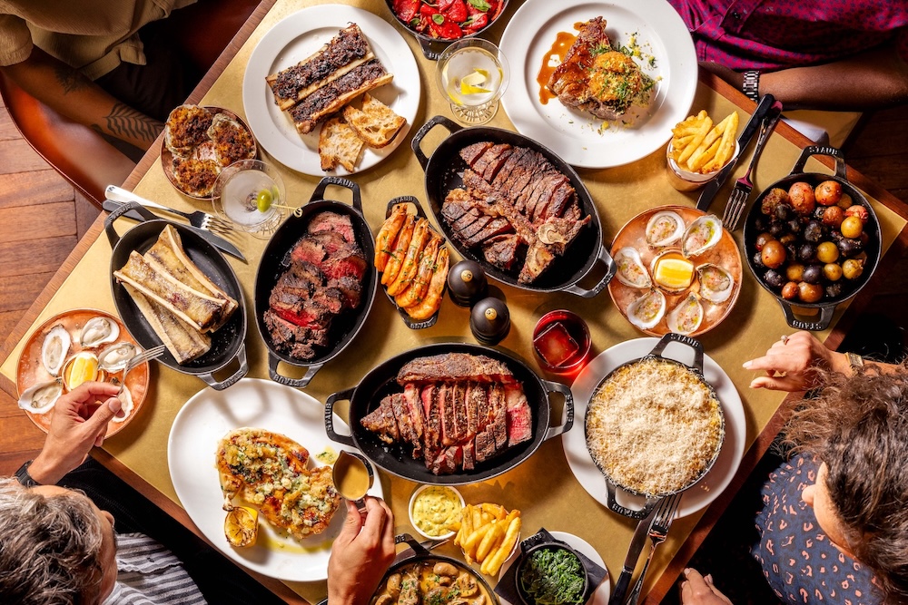 An above view of people sat at a table that is full of sharing steak in black skillets, sides, such as fries and mac and cheese and cocktails.