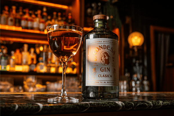 A bottle of Condesa Gin Clásica with a wooden cap stands on a marble bar counter next to a filled stemmed cocktail glass, with a softly lit, out-of-focus bar and liquor shelves glowing warm orange in the background.