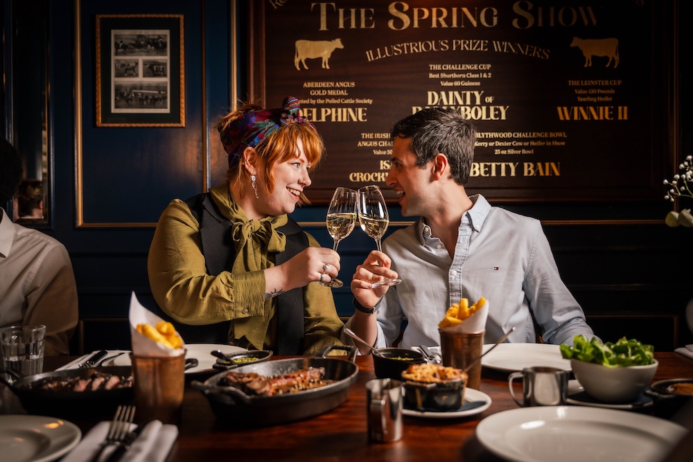 A couple sat at a dining table, looking at each other and clinking wine glasses. On the table in front of them is a black skillet of sharing steak, two portions of fries and mac and cheese.