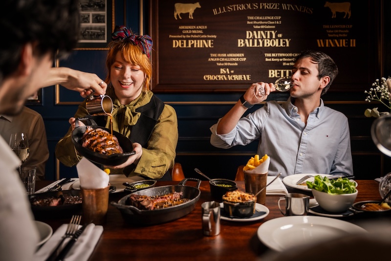 Three people at the dining table, one of the men is drinking wine, whilst the woman is holding a skillet of sharing steak. Another man is pouring sauce over the steak.