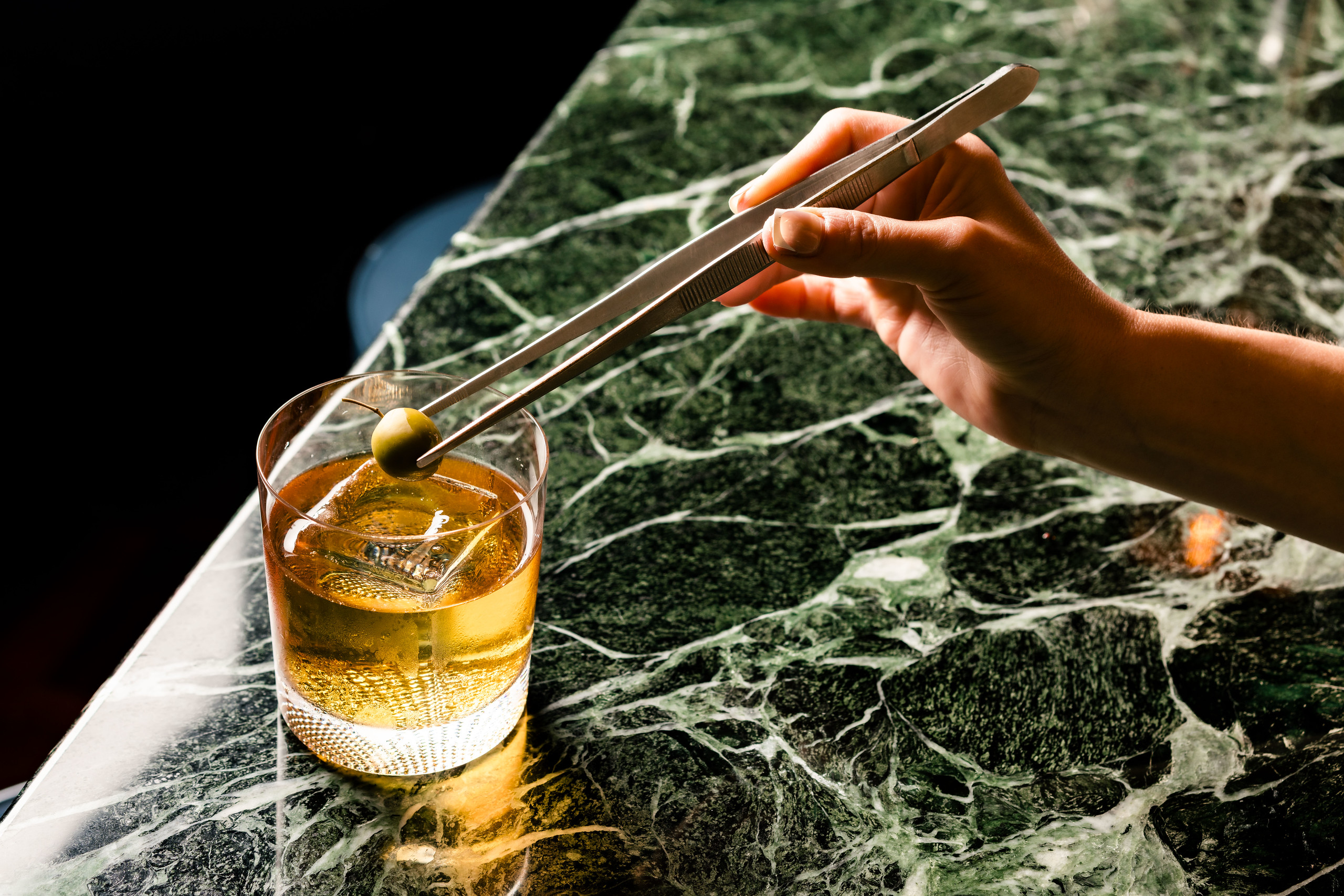 light amber cocktail placed on the edge of a bar, a hand holding tweezers carefully placing an olive in the glass as a garnish