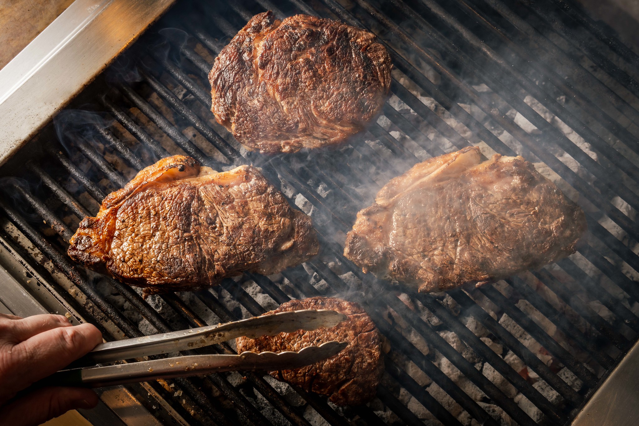 A chef turning steaks with tongs over the charcoal grill at Hawksmoor, smoke rising from the bars