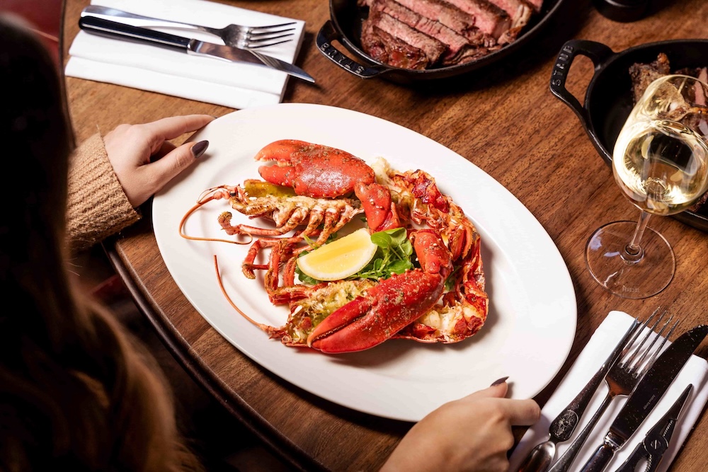 A whole lobster placed on a table in front of a guest. In the centre of the table are two skillets of sharing steak and a glass of white wine.