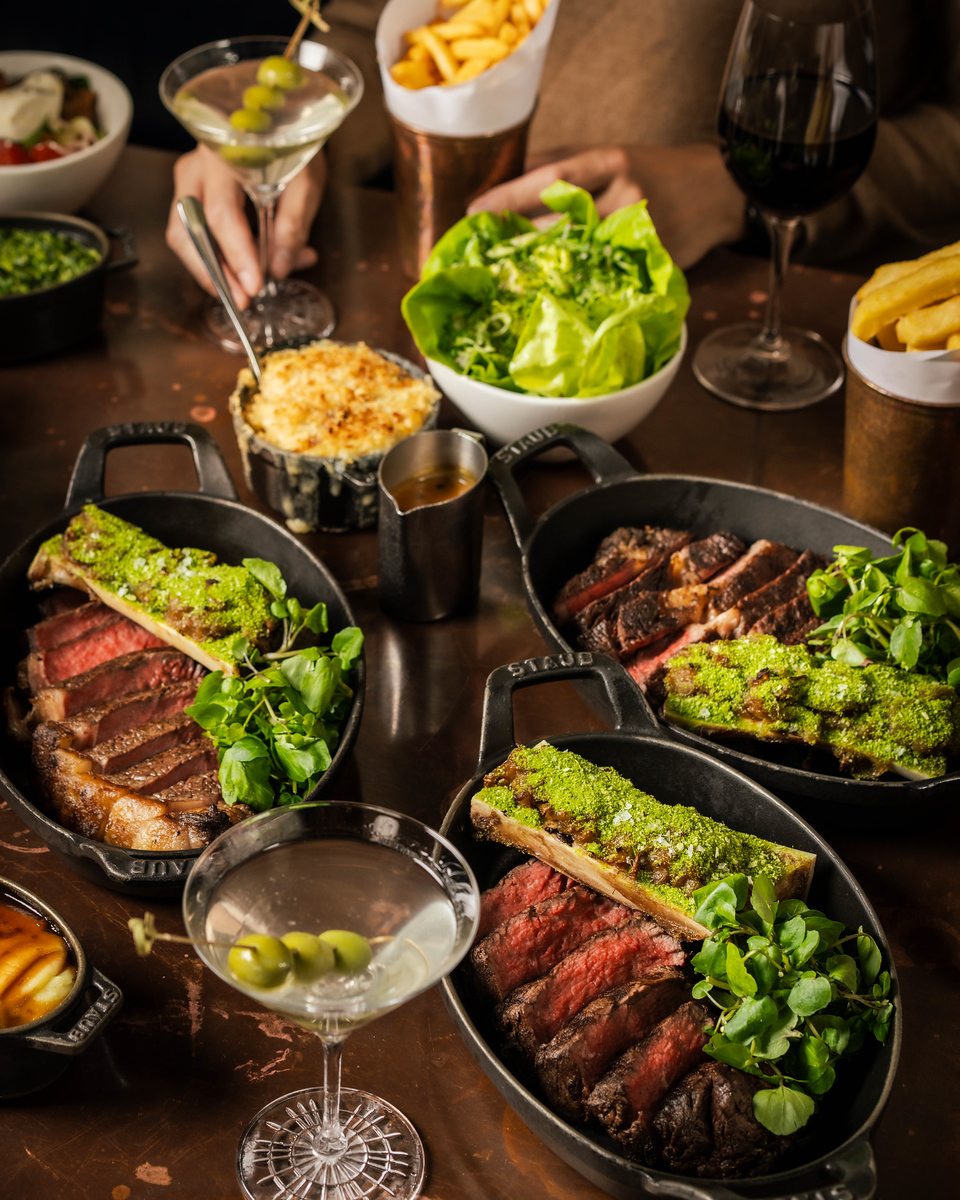 A Hawksmoor steak flight on the table, sliced steaks in cast iron dishes topped with marrow bone and watercress, with sides and martinis
