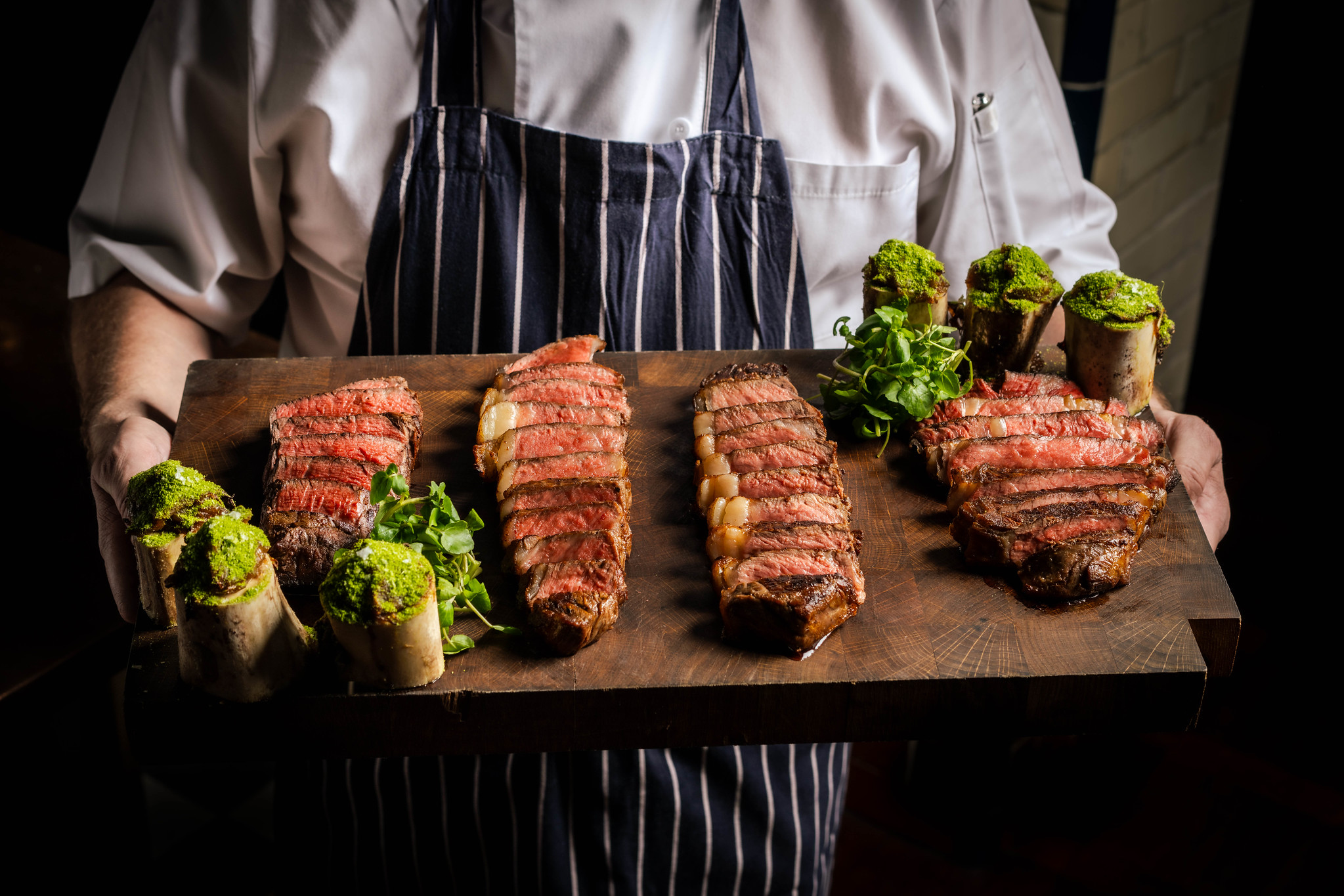 A Hawksmoor chef carrying the steak flight to the table, four cuts sliced on a wooden board with greens