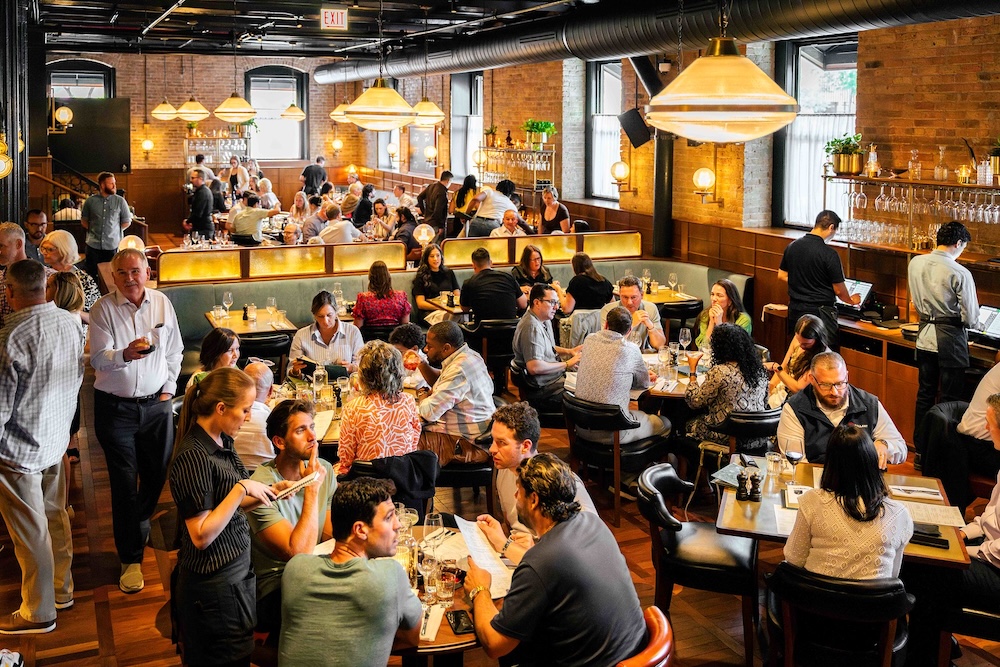 A bustling lunch time at Hawksmoor Chicago - the dining room is filled with guests sat at tables. Low hanging lights illuminate the room with a yellowish tinge.