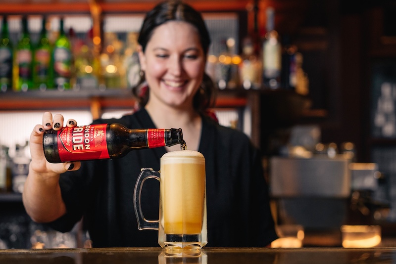 A bottle London Pride being poured into a beer mug by a smiling female bar tender. The beer is foaming up as it is being poured, forming a thick head at the top of the mug.