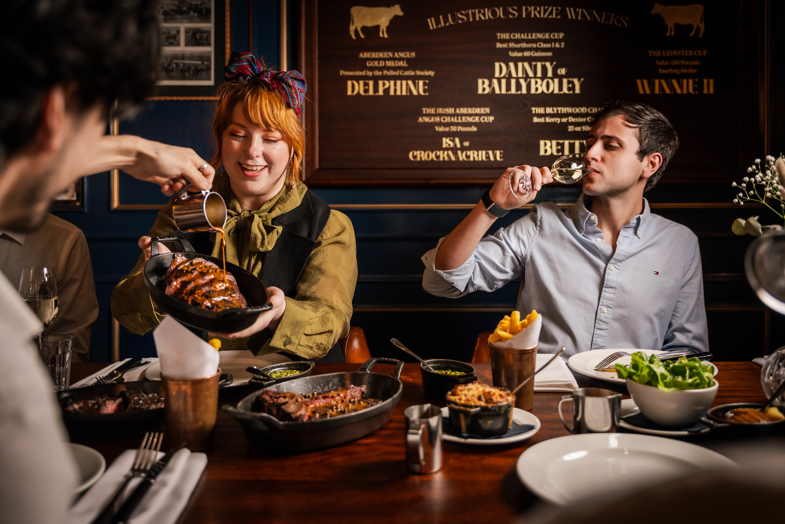 A couple at the dining table, the man is drinking wine, the woman is holding a skiller of sharing steak