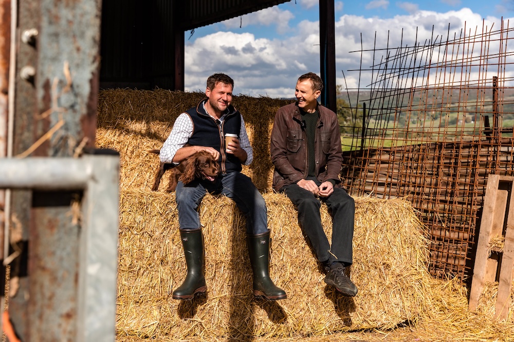 Two men, including the Hawksmoor founder Huw, sitting on a hay bale, smiling and chatting in a barn with no walls. Huw is holding a takeaway cup of coffee in his left hand and stroking a young brown Springer Spaniel with his right.