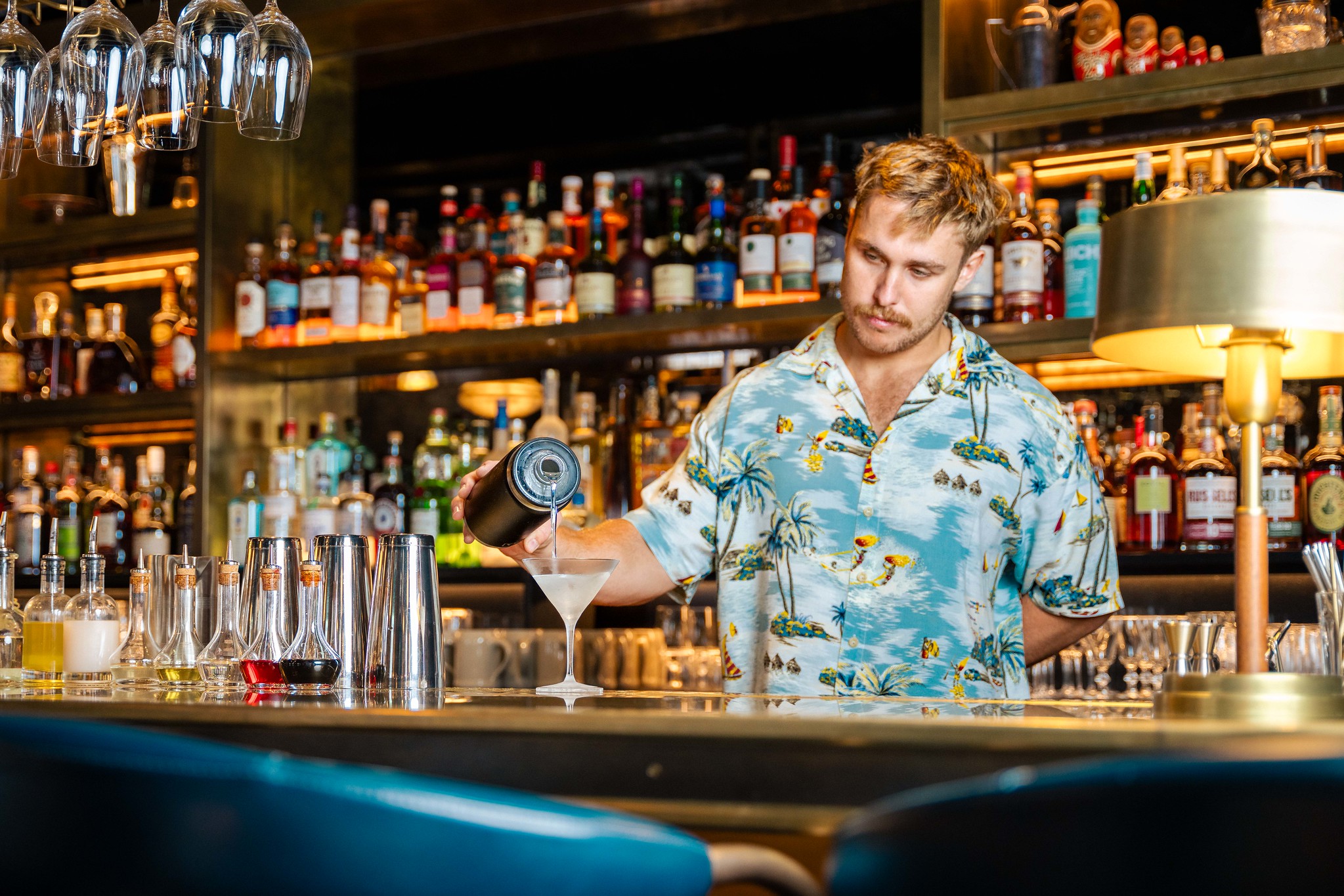 A casulally dressed bartender in a light blue shirt bvehind the bar, pouring cocktails