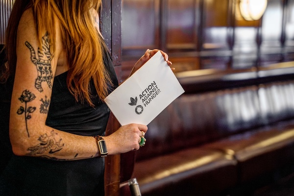 A woman with tattoos on the arm holding a card with the 'Action Against Hunger' logo on the back