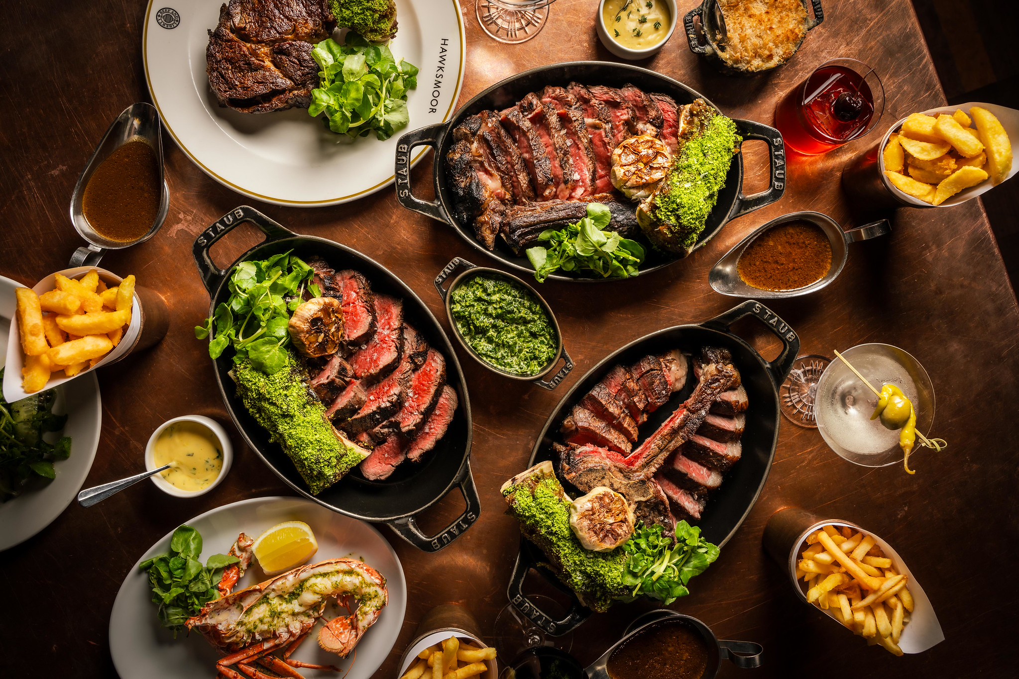 Overhead spread of sliced steaks in cast-iron dishes with fries, lobster, watercress, sauces and cocktails on a wooden table