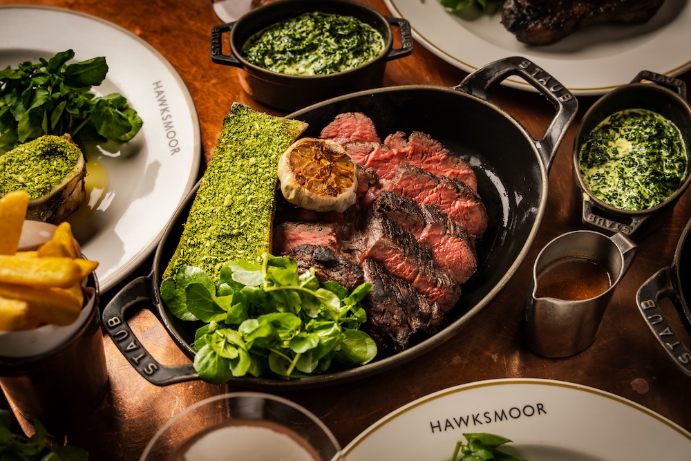 A sharing steak placed in the centre of the table, presented in a black skillet with a jug of bone marrow broth besides it. The edges of three 