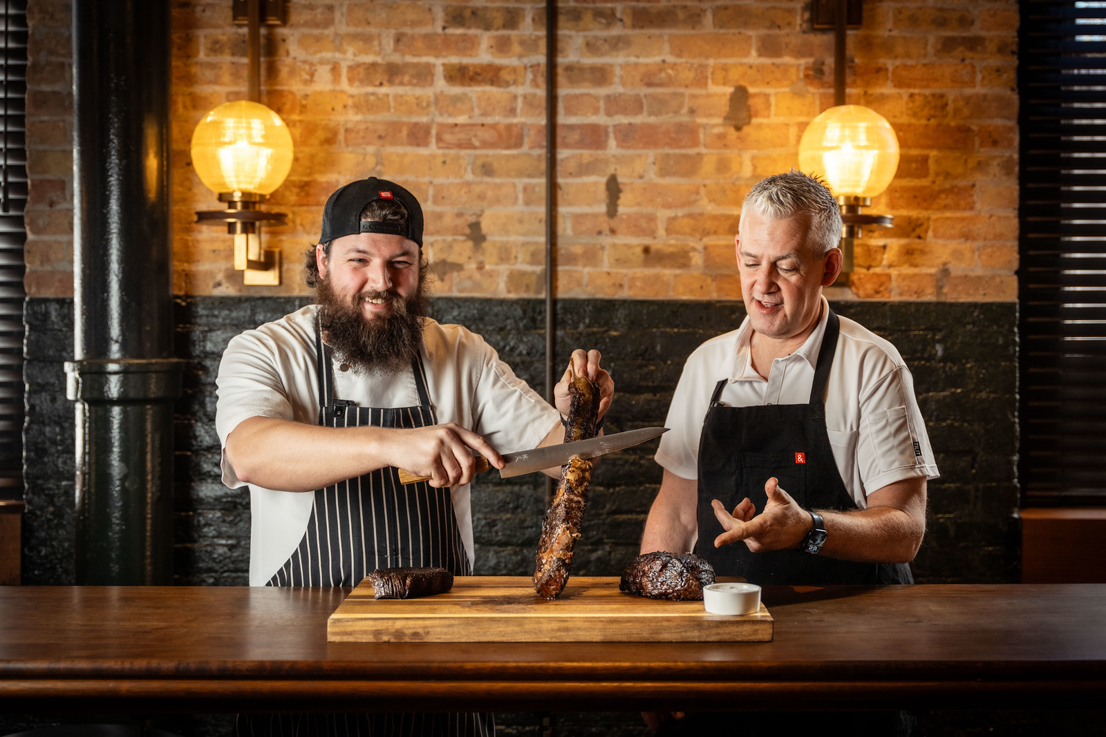 Two chefs behind a table, smiling at the camera; one of them of holding up a meat skewer and slicing a piece off it