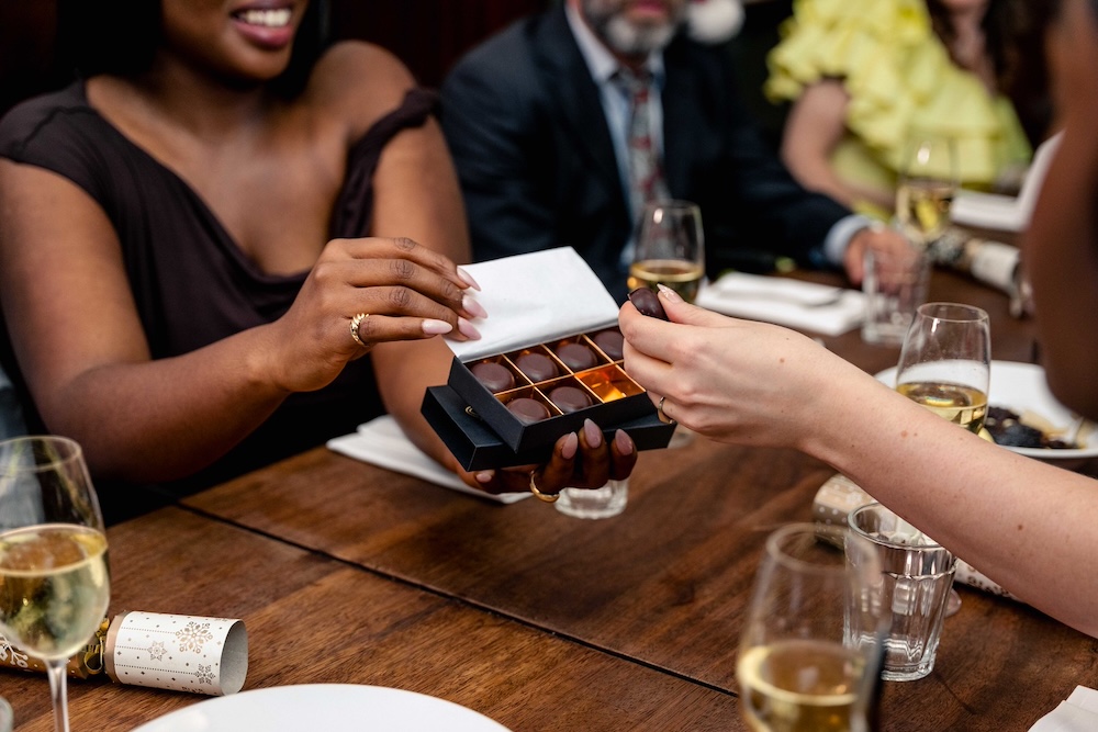 A smartly dressed woman in a black dress smiling whilst holding up a box of Trubutes chocolates, another woman's hand is pictured picking up one chocolate.