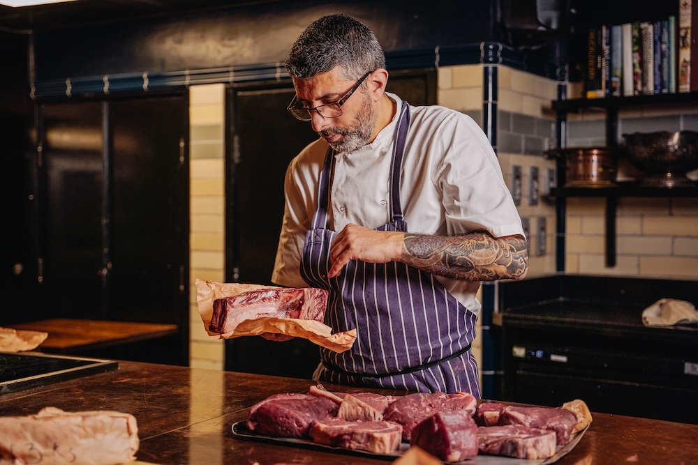 A chef, in chefs whites and a blue apron inspecting different cuts of steaks laid out on the table in front of him in a kitchen.