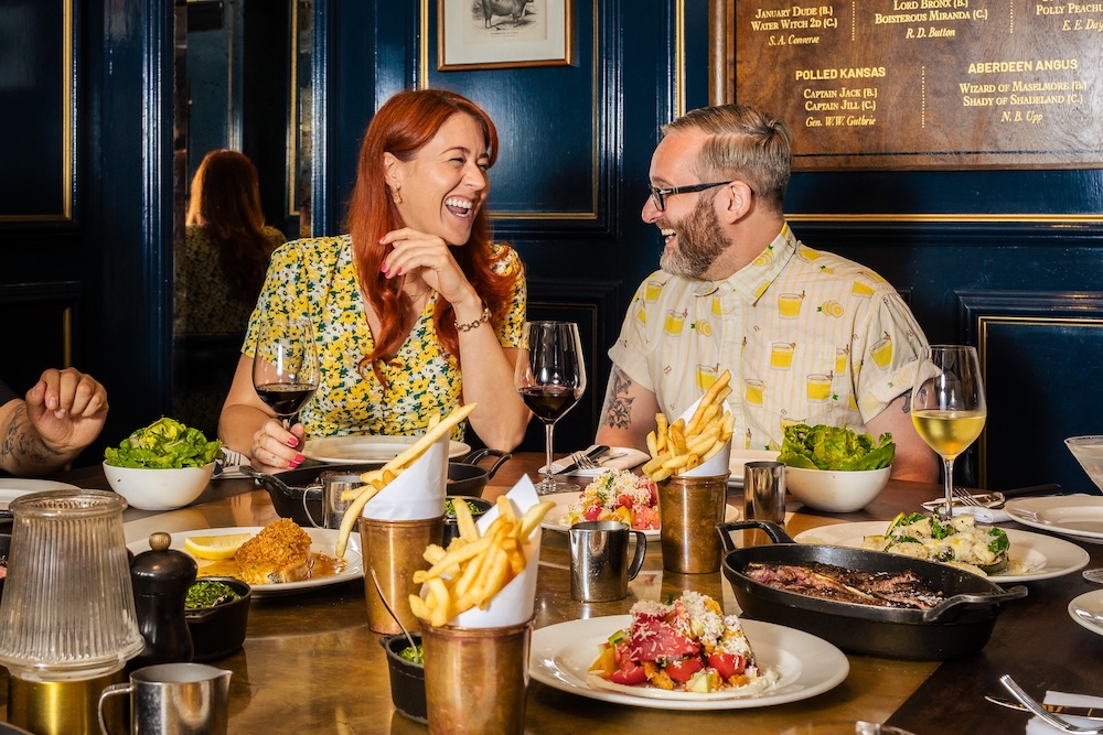 A man and woman sat laughing to one another whilst sitting at a table full of food, including a sharing steak in a black skillet, fries and wine.