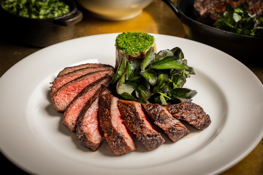 Placed on a shiny golden coloured table is a Picanha wagyu steak cut into thin slices served on a pristine white plate, resting against bone marrow. In the background a dish of creamed cabbage is visible.