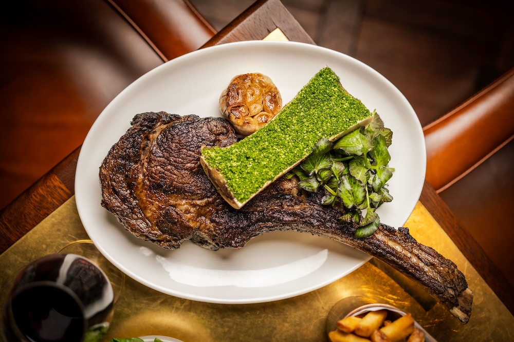 An above view of a long bone rib chop on a white plate with a side of bone marrow with green garlic-parsley crumb on a table. Next to it is a glass of red wine and side of fries.
