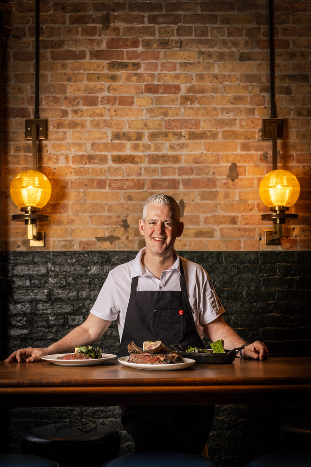 A smiling chef behind a table with three large plates of sliced steak. There is a brick wall behind him with two large circular lights attached to it, giving of a gentle yellow glow.