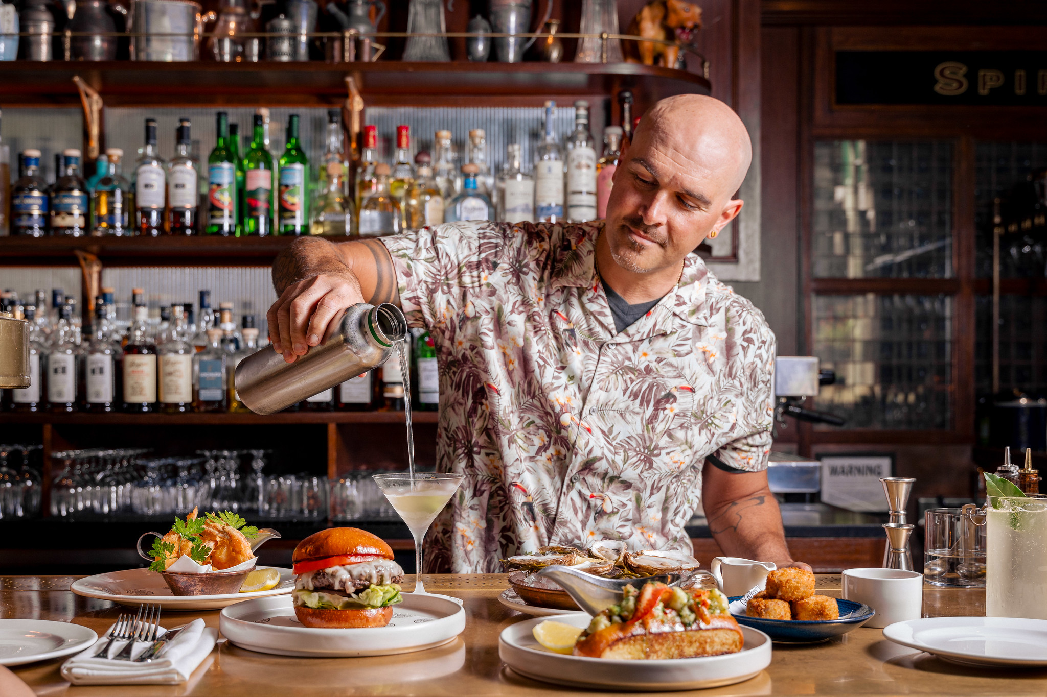 A casually dressed smiling bartender pouring a cocktail behind the bar