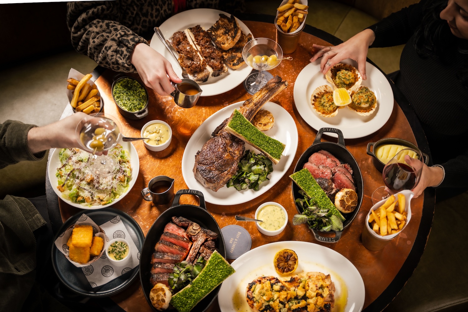 An overhead shot of a circular wooden table laden with cuts of steak in skillets, sides including fries and drinks of martinis and red wine. The arms of four guests are seen picking up food items.