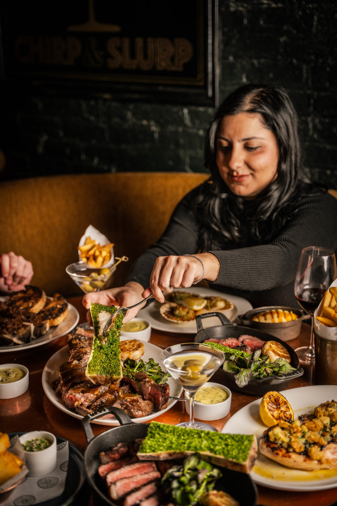 A young attractive woman scooping some bone marrow onto a steak. The table around it is generously laden with steaks, sideas and drinks