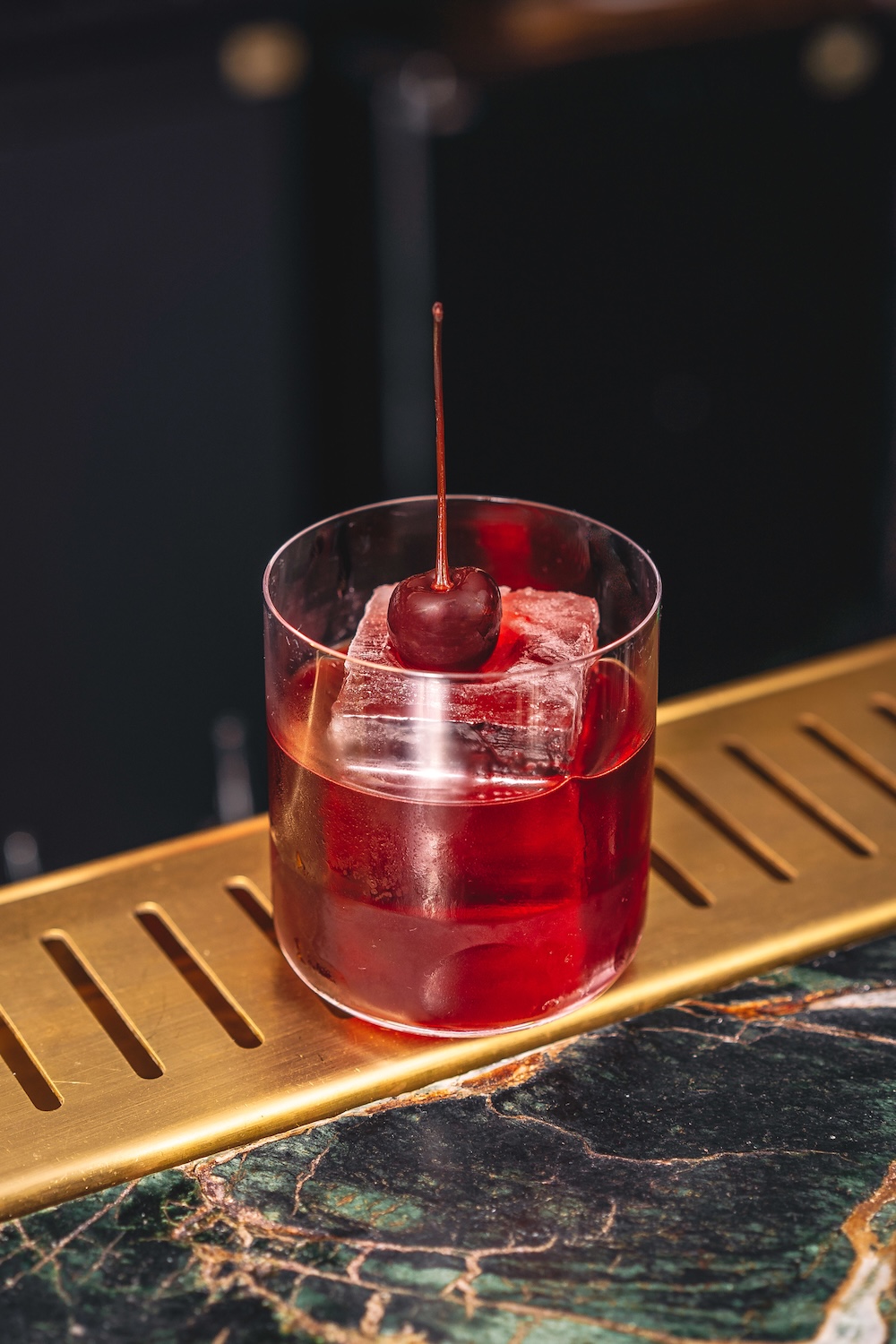 A cherry negroni cocktail, in a glass with a large ice cube in it and garnished with a cherry.