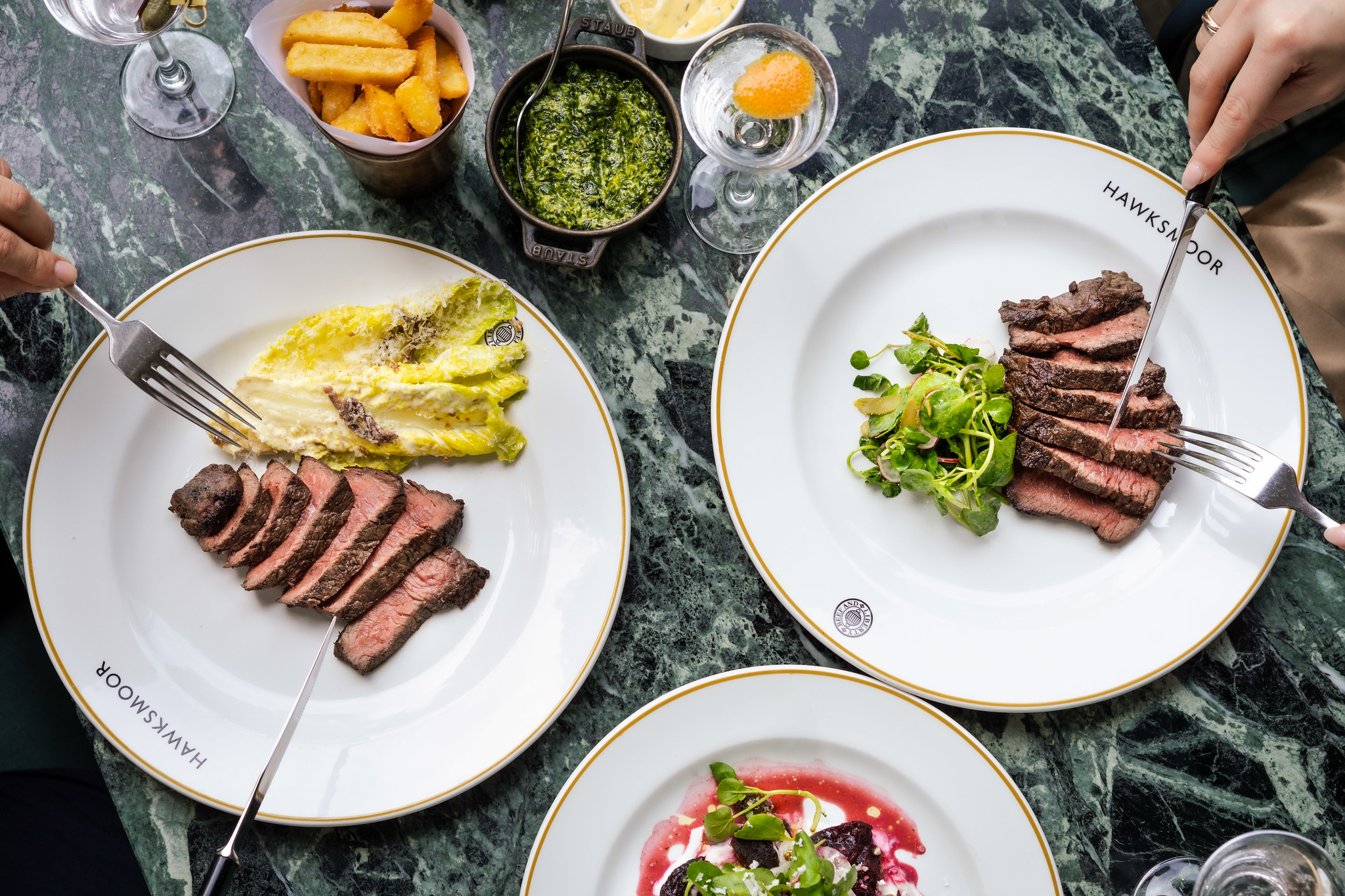 An overhead shot of a marble table with two white plates, with sliced steak and salad