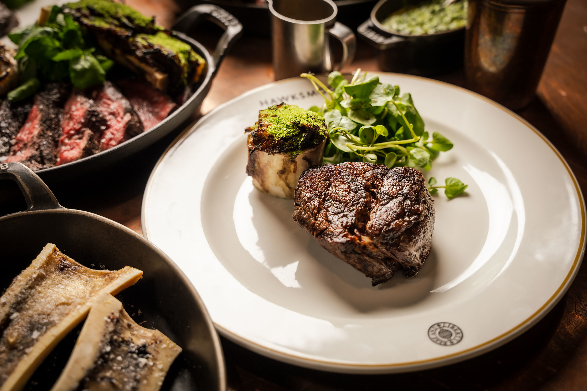 Thick-cut steak on a white plate with herb-crusted bone marrow and watercress, with more steak behind