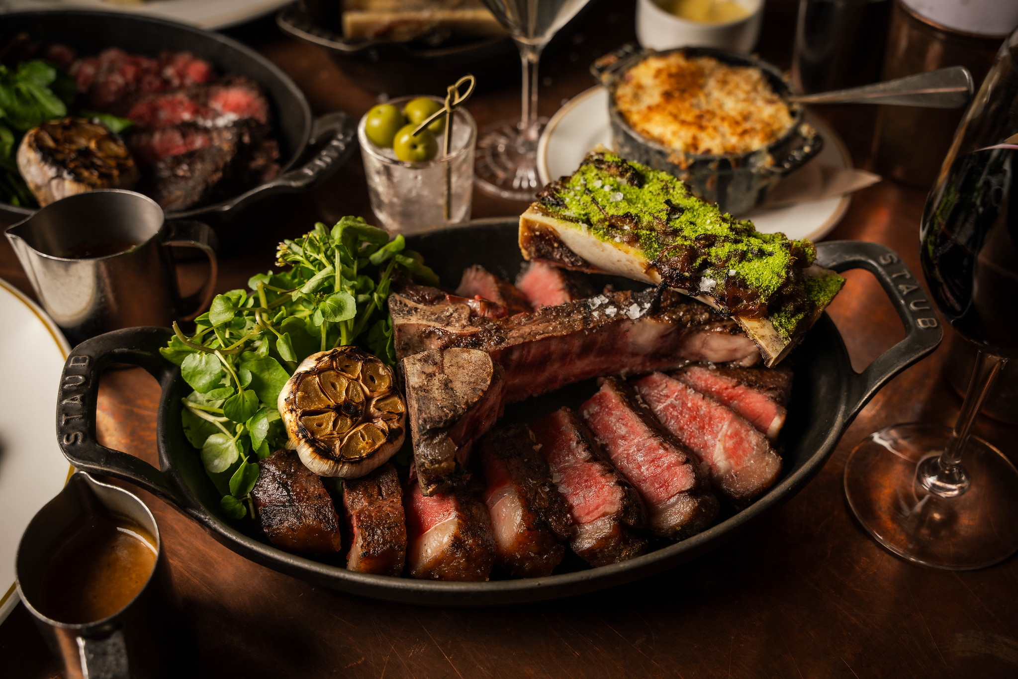 Sliced bone-in steak in a cast-iron dish with herb-crusted bone marrow, roasted garlic and watercress