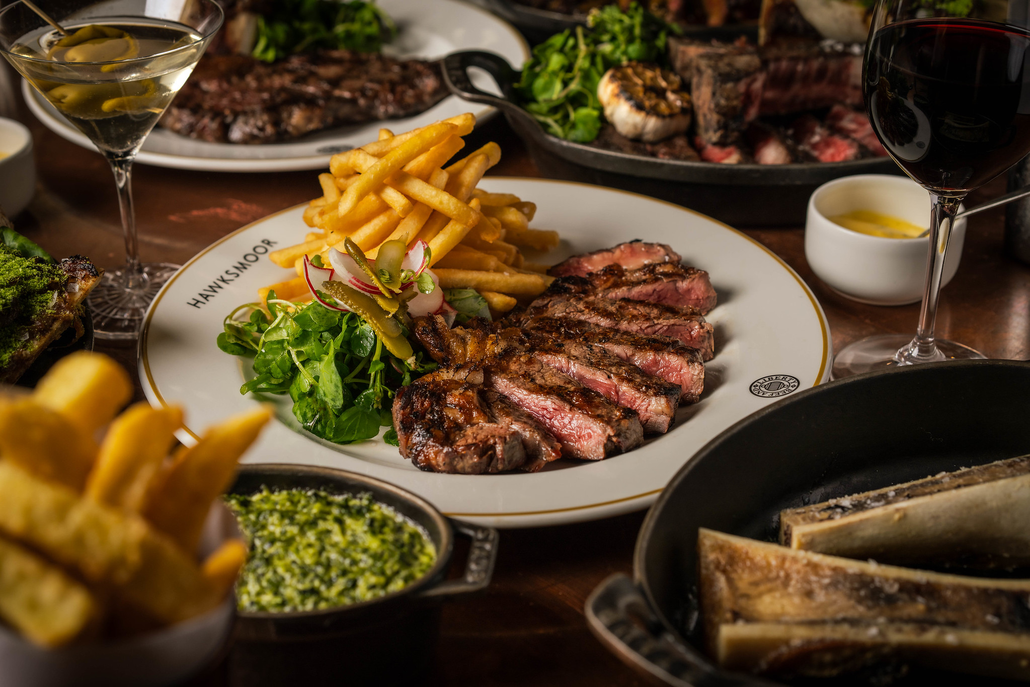 Sliced grilled steak on a white plate with fries and watercress, with bone marrow and wine nearby