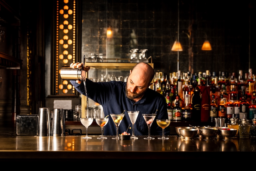Drinks guru Liam, dressed in a smart black shirt, pouring a line up of six martinis at the bar