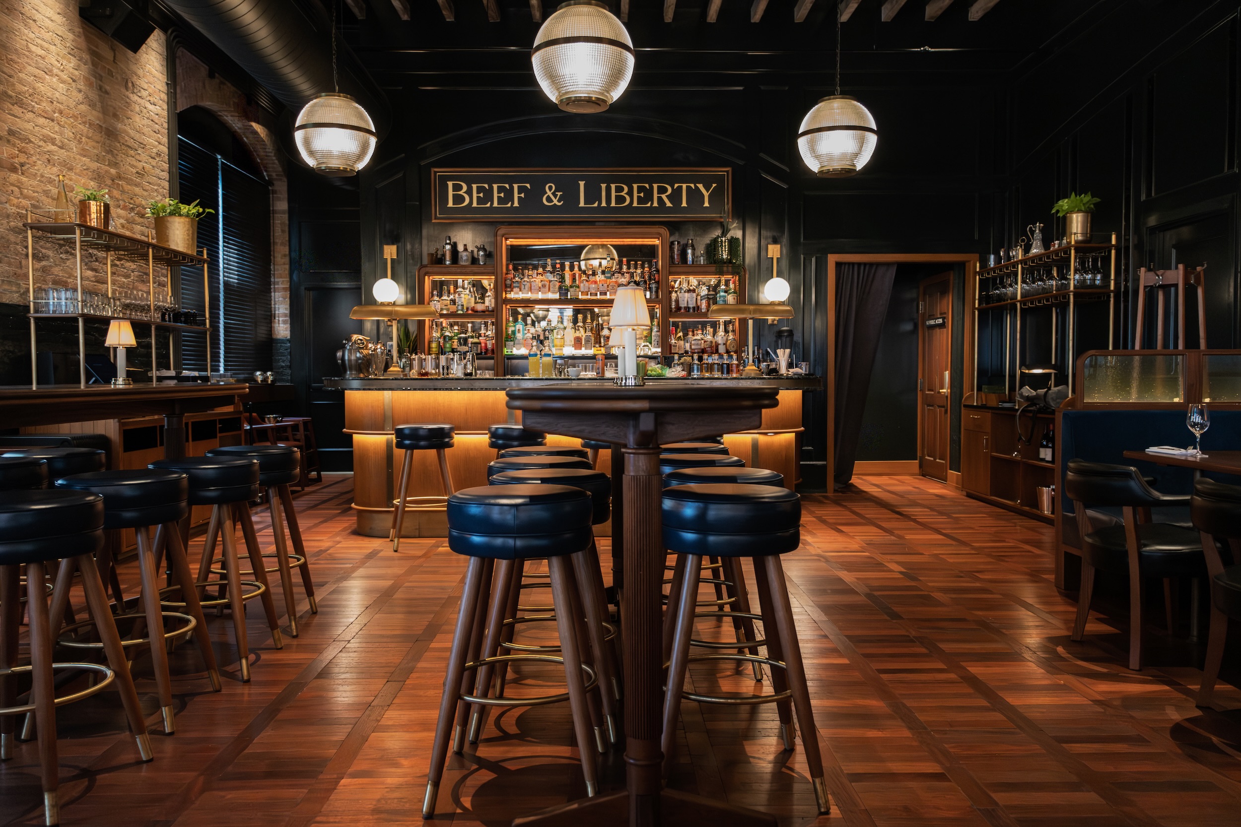 The "Beef & Liberty" bar area featuring blue stools around a marble counter, wood tables, small lamp lights, and a backdrop of spirits and glassware.