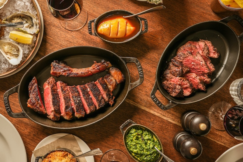 An above view of two large Hawksmoor sharing steaks served in black skillets, on a wooden table. There are sides of oysters, creamed spinach and mac and cheese placed around the steaks.
