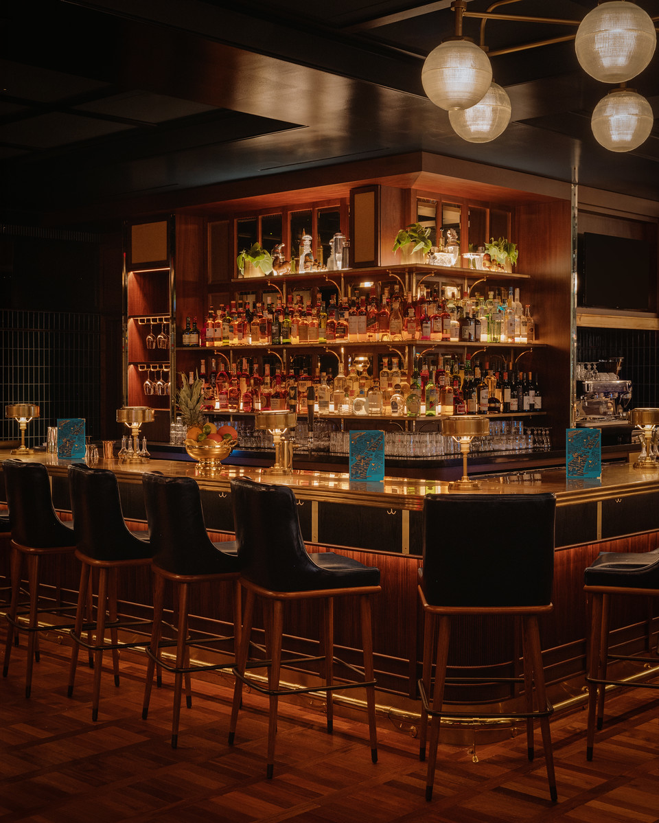 The bar at Hawksmoor Boston: a curved counter with a brass rail and wooden stools, cocktails set along it, tiered shelves of backlit bottles behind, and globe pendant lights overhead.
