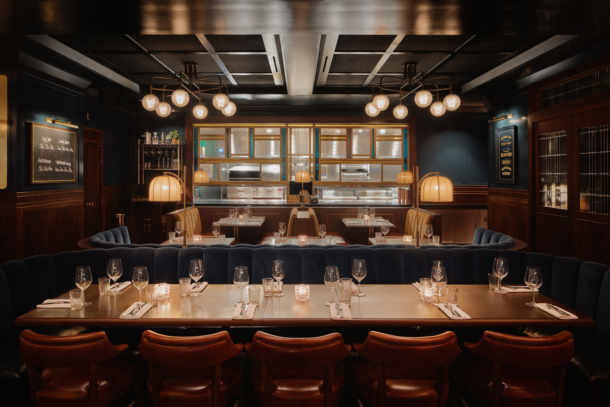 Restaurant interior at Hawksmoor Boston: a long counter set with glassware and lined with tan leather stools, dark wood panelling and deep blue walls, and backlit shelves beneath clustered globe pendant lights.