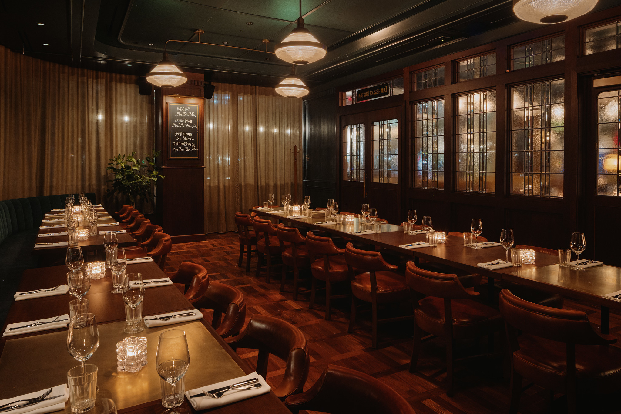 The Duchess private dining room at Hawksmoor Boston: a long table laid with glassware and napkins, tan leather chairs down both sides, dark panelled walls with glass-fronted cabinets, and dome pendant lights overhead.