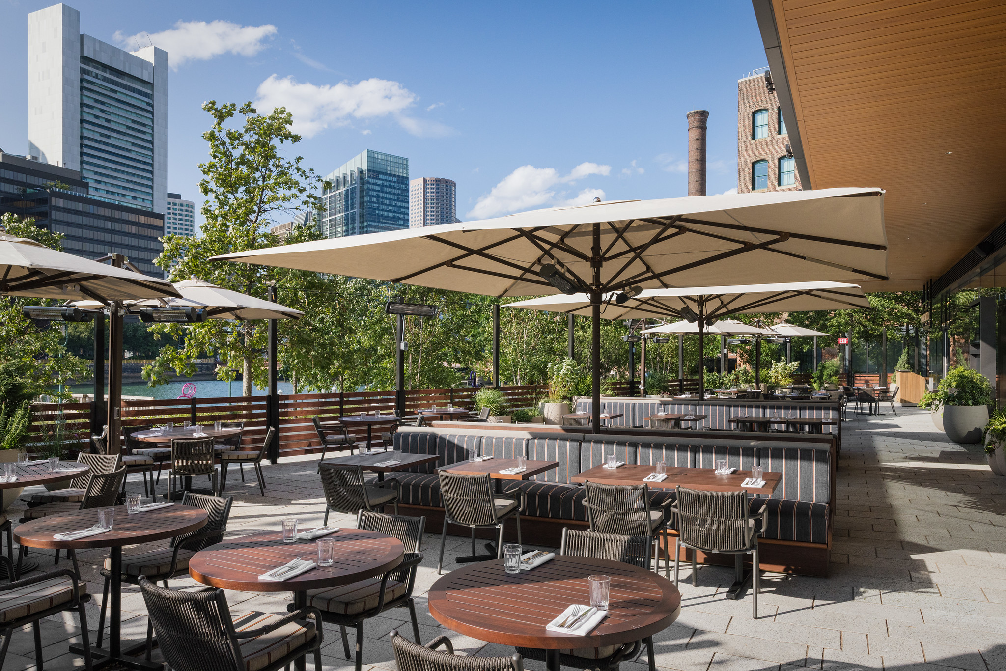 The terrace at Hawksmoor Boston on a clear day: wooden tables and chairs under cream parasols, planting along the edge, and glass towers rising behind.