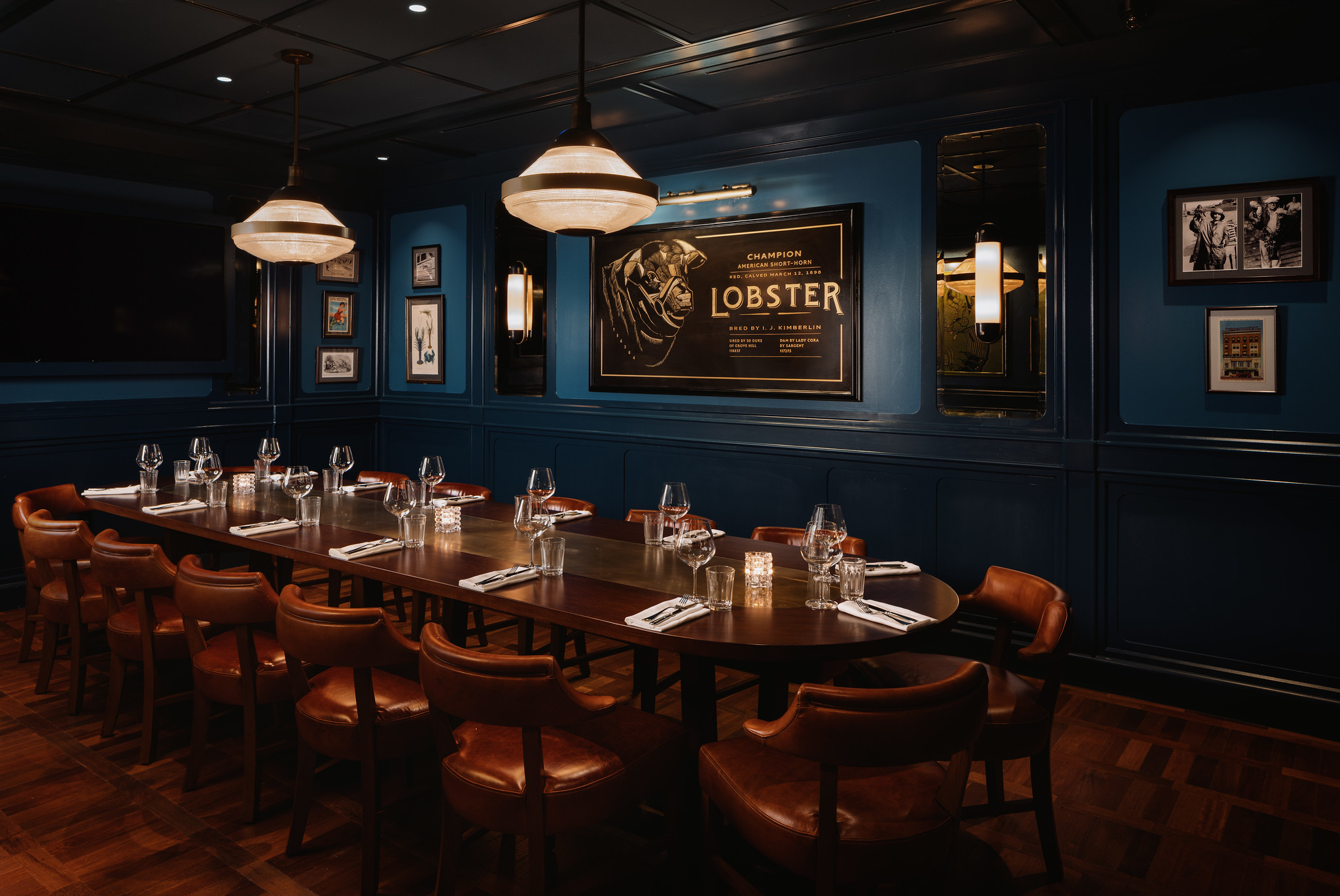 The Lobster private dining room at Hawksmoor Boston: one long table laid with glassware and candles, tan leather chairs down both sides, deep blue panelled walls hung with framed pictures, and dome pendant lights overhead.