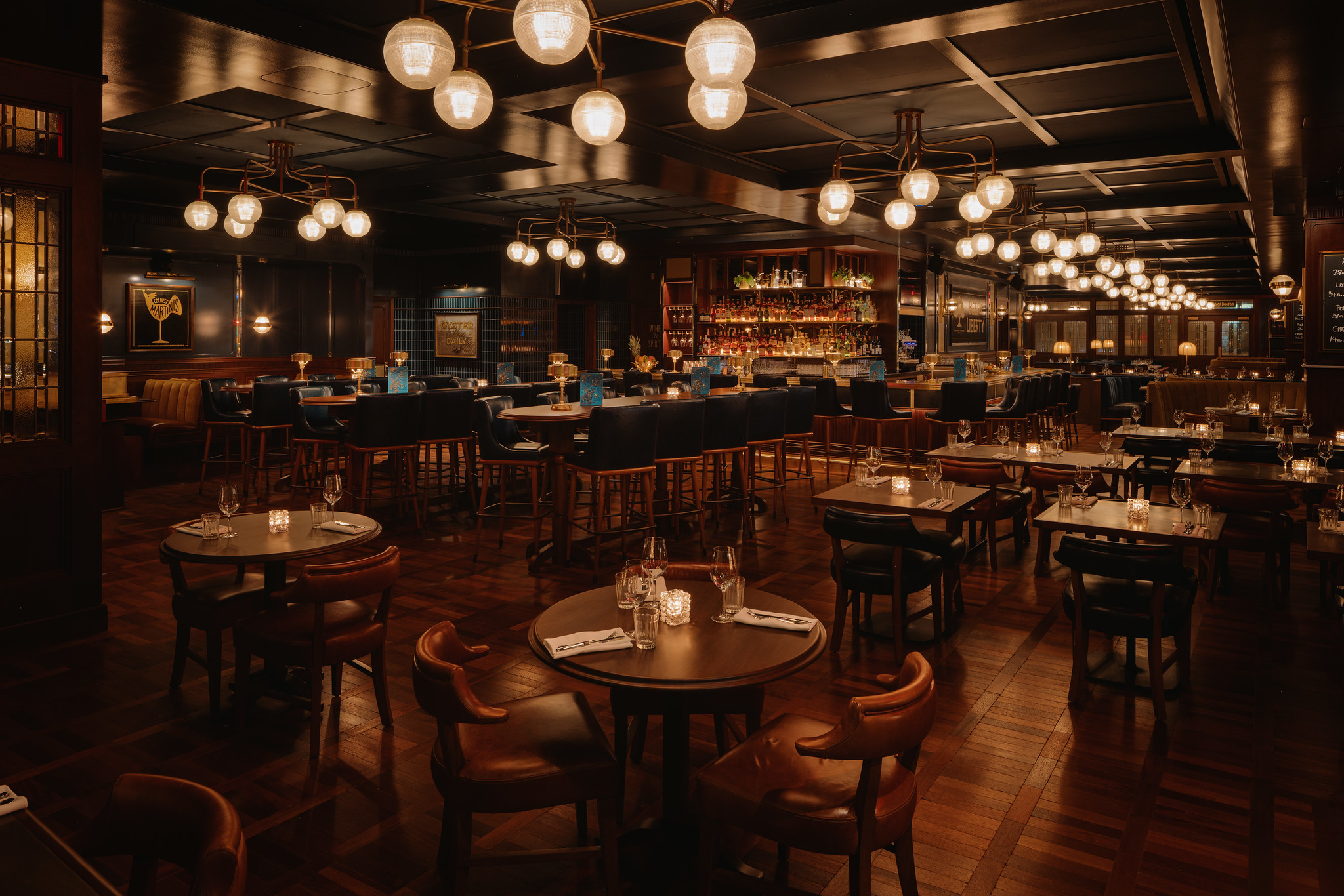 The main dining room at Hawksmoor Boston: rows of wooden tables and chairs on a parquet floor, and clusters of globe pendant lights hung at different heights above them.
