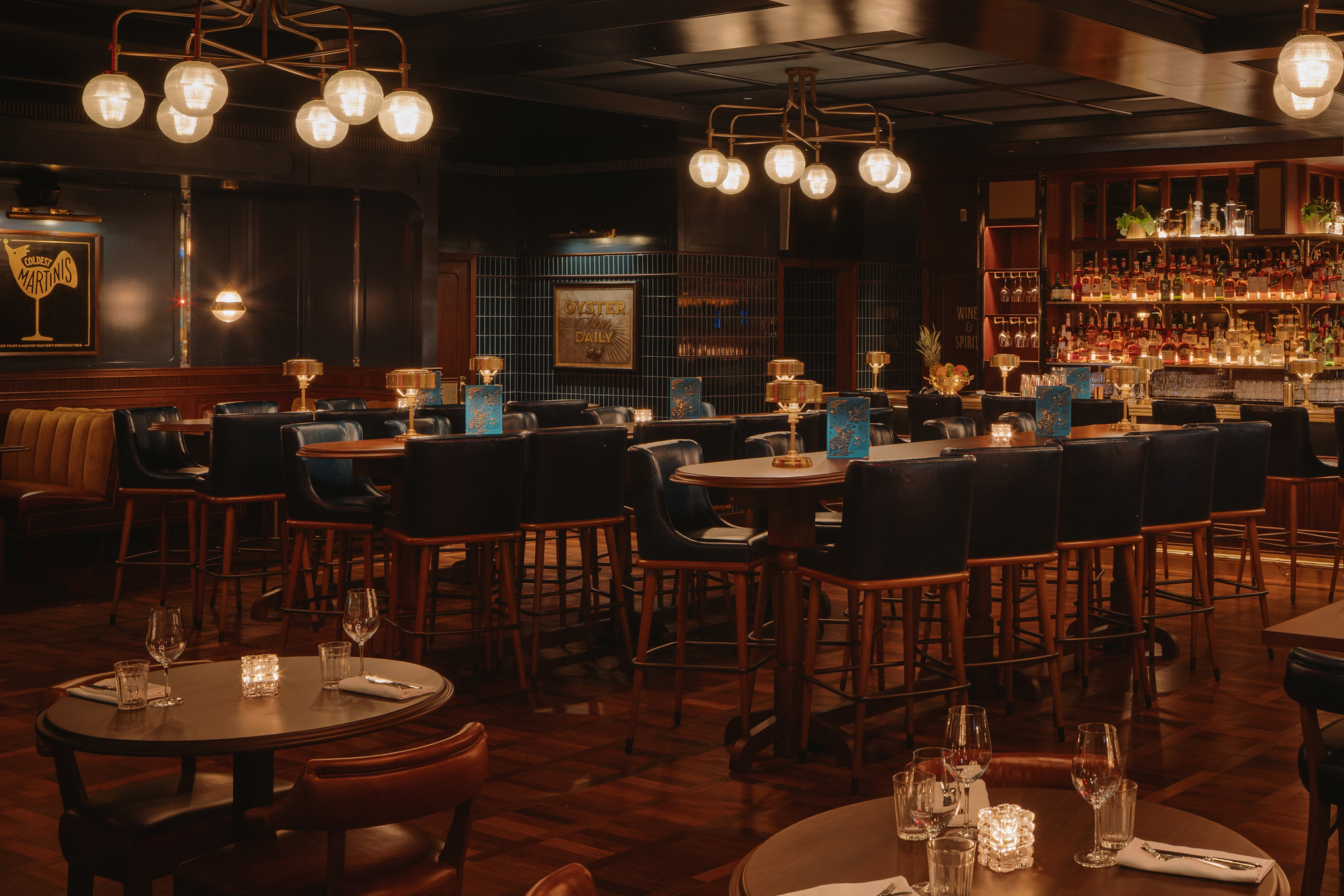 High top tables and stools in the bar at Hawksmoor Boston, with globe pendant lights and a back bar lined with bottles
