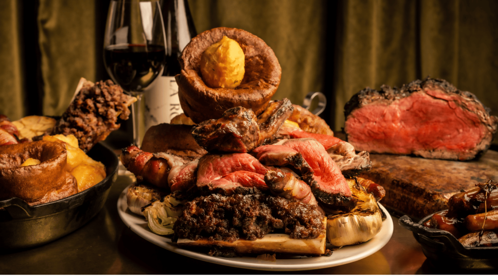 A large pile of roast beef on a plate, topped with Yorkshire puddings on a dark brown wooden table.