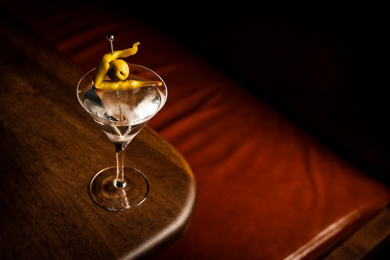 A martini served in a classic martini glass that is garnished with an olive and green chilli, placed on a dark wooden table.