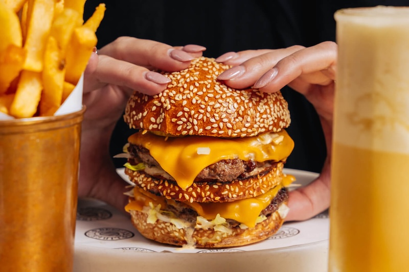Hawksmoor Chicago Notorious Beef Burger - pictured closely, being picked up by two hands, with a serving of fries and a cocktail in the foreground.